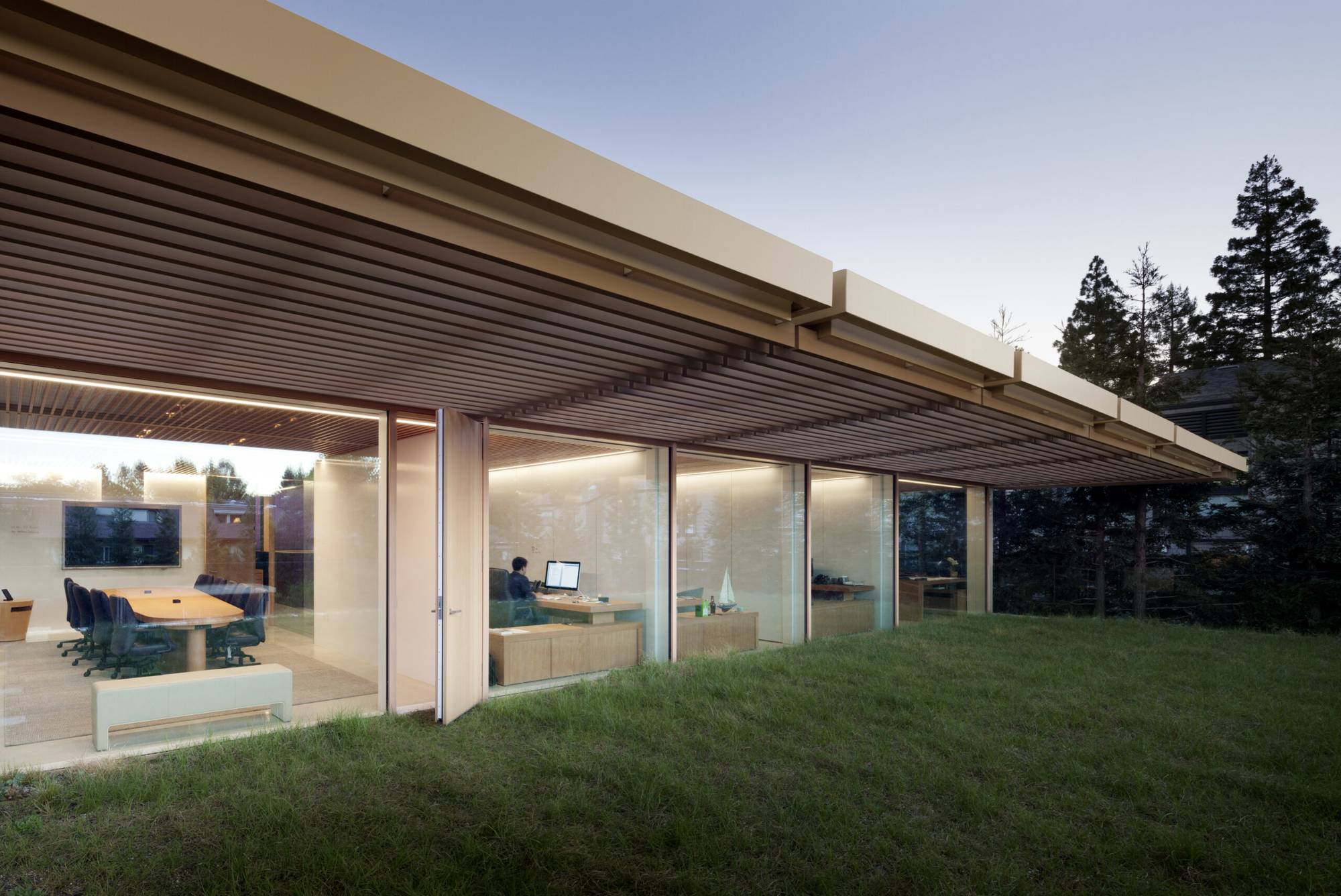 A glass-walled office opening onto a lawn at dusk, with a meeting room and a person working at a desk
